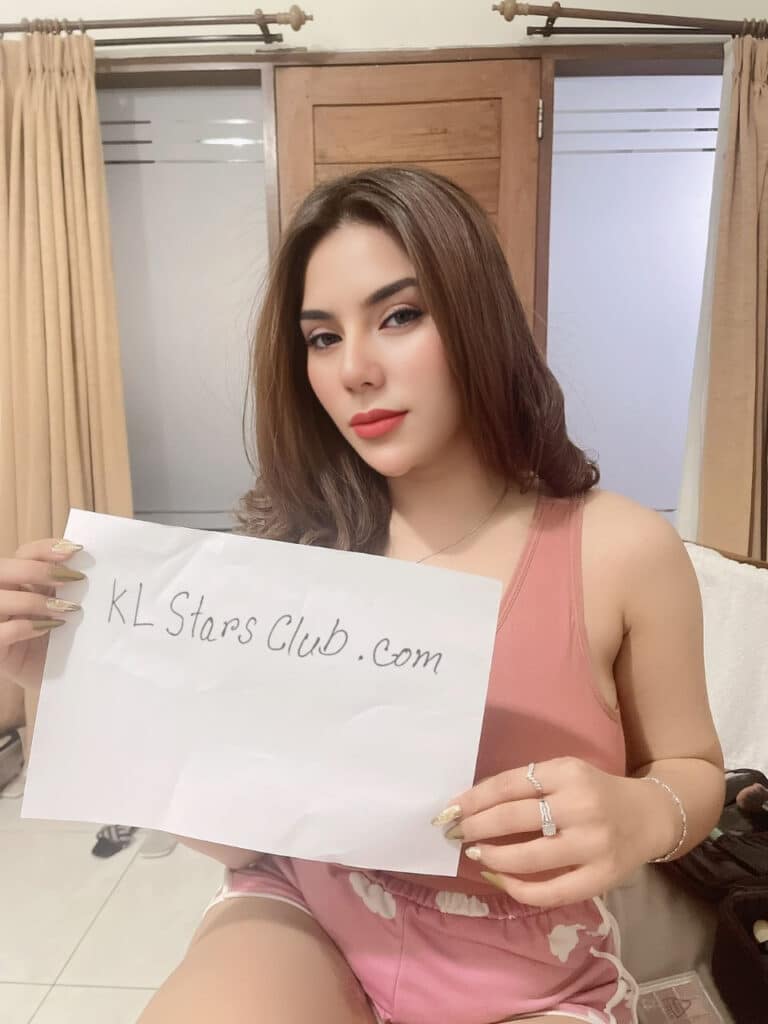 S-class Kuala Lumpur escort Victoria Qistina holding a sign for KL Stars Club in a cozy indoor setting, wearing a pink sleeveless top and shorts.
